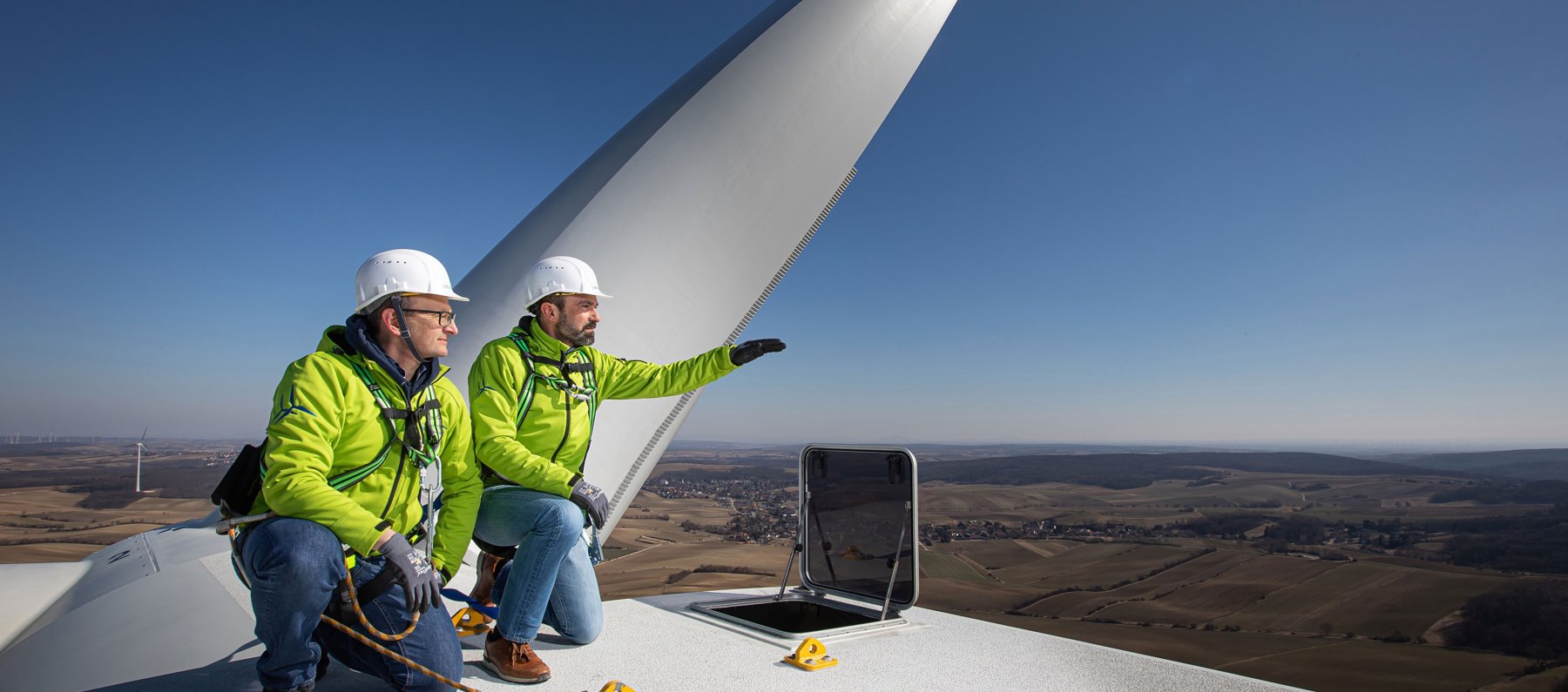 Die Geschäftsführer der Windkraft Simonsfeld auf einer Windkraftanlage. Über Ihnen der blaue Himmel. Hinter Ihnen ein Flügel der Windkraftanlage.